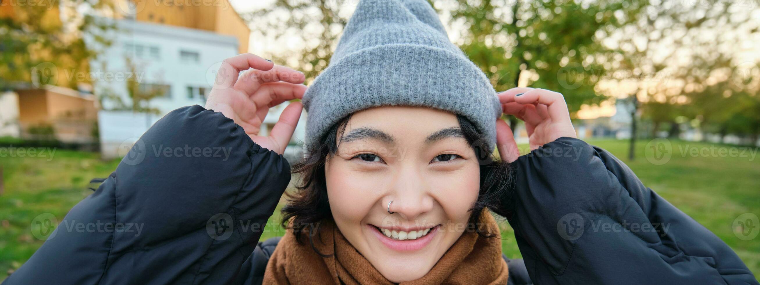 Portrait of cute asian girl in hat and scarf, walks around town in chilly spring weather, smiles and looks happy, sits in park near green grass photo