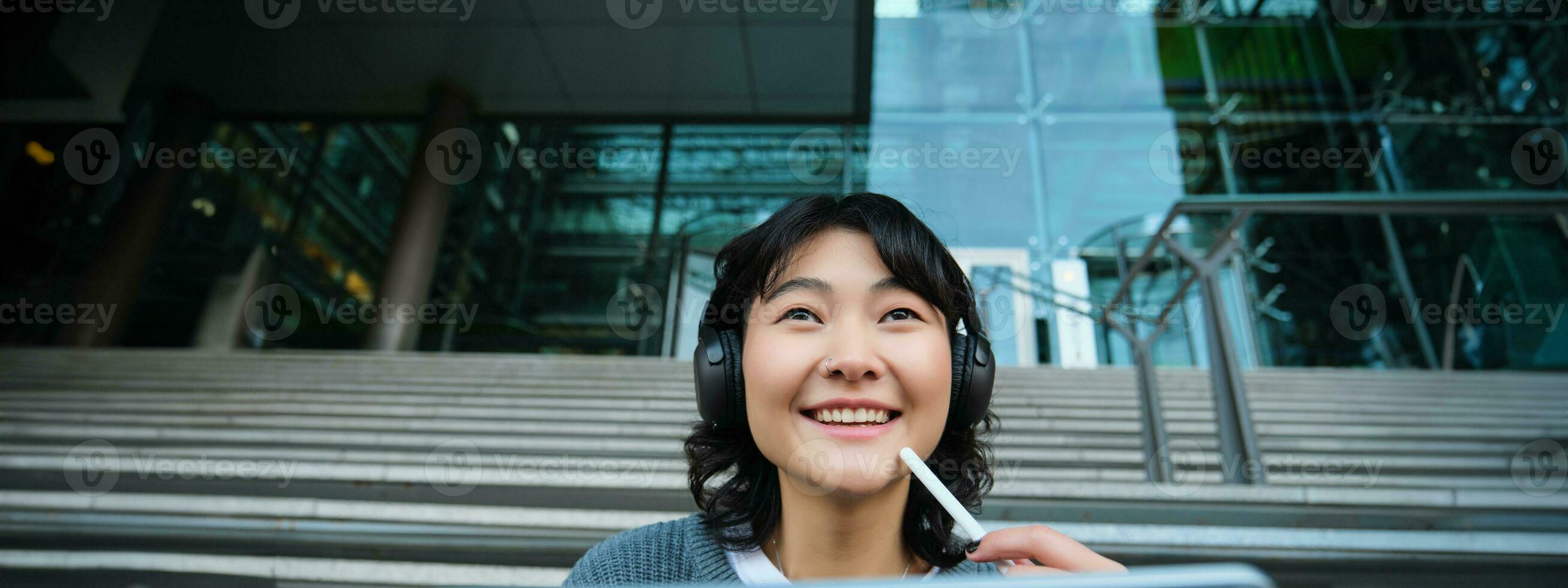Close up of smiling asian girl feels inspired while draws on digital tablet, makes graphic design project, pencils user interface assignment, sits on stairs near campus photo