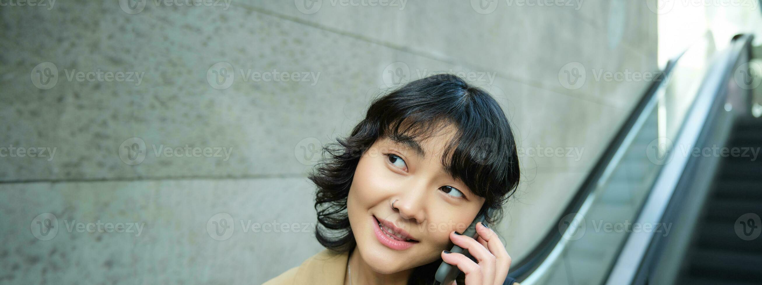 Close up portrait of smiling brunette girl, korean woman goes down escalator, makes phone call, talks to someone on telephone photo