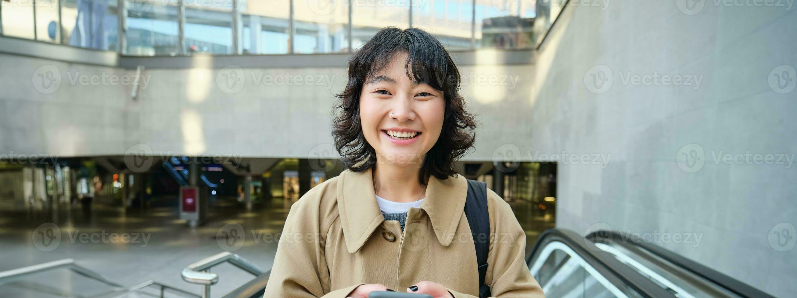 personas en ciudad. joven coreano mujer viajes alrededor ciudad, va arriba el escalera mecánica, usos su móvil teléfono y sonrisas foto