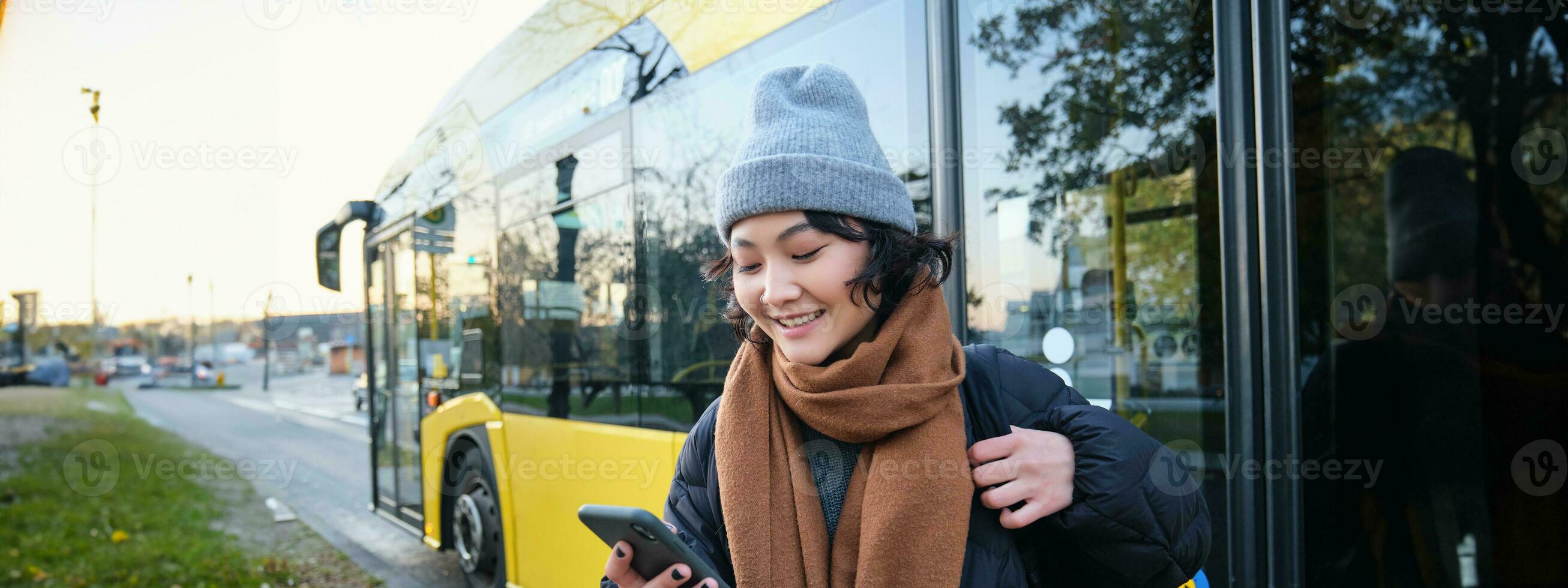 Image of girl student waiting for public transport, checks schedule on smartphone app, stands near city bus photo