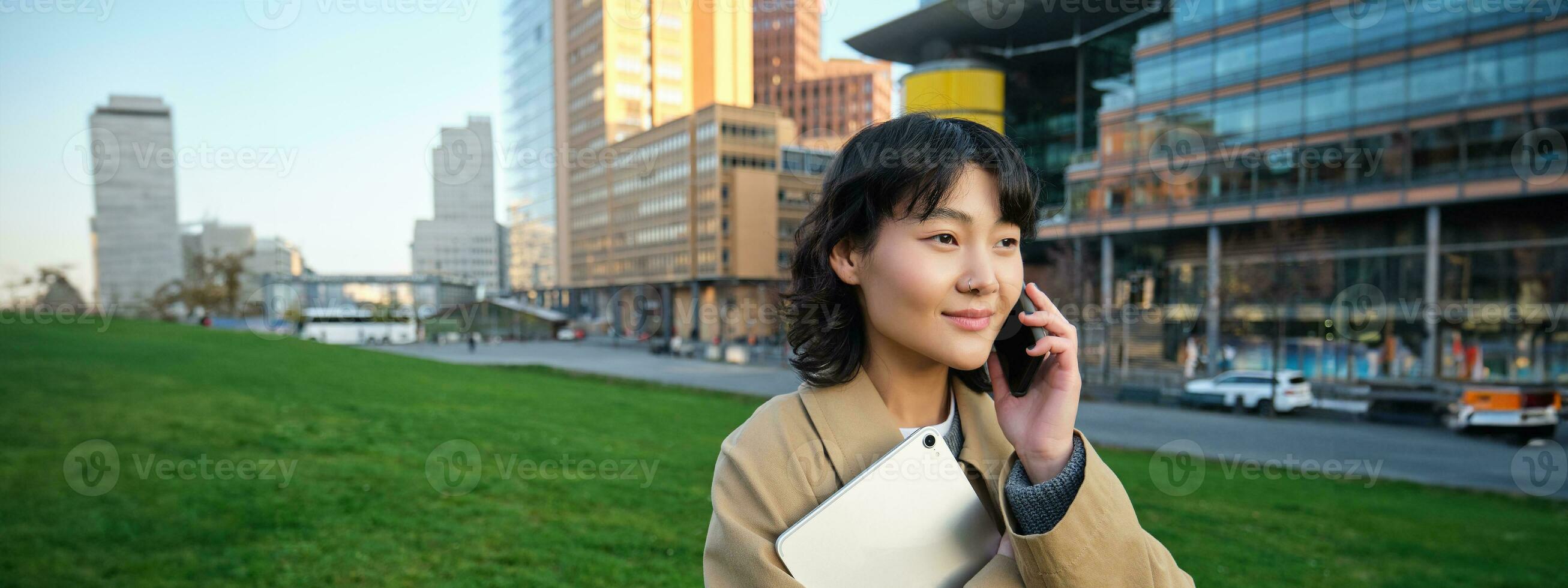 Stylish smiling girl, university student in trench coat, holds tablet, talks on mobile phone, has conversation over telephone and looks relaxed, stands on street photo