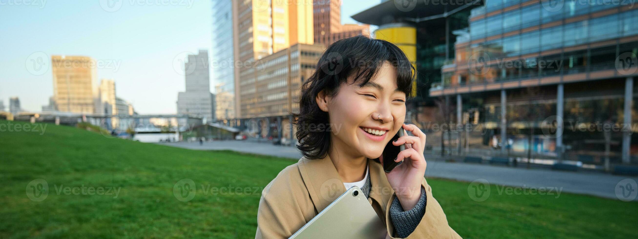 Stylish smiling girl, university student in trench coat, holds tablet, talks on mobile phone, has conversation over telephone and looks relaxed, stands on street photo