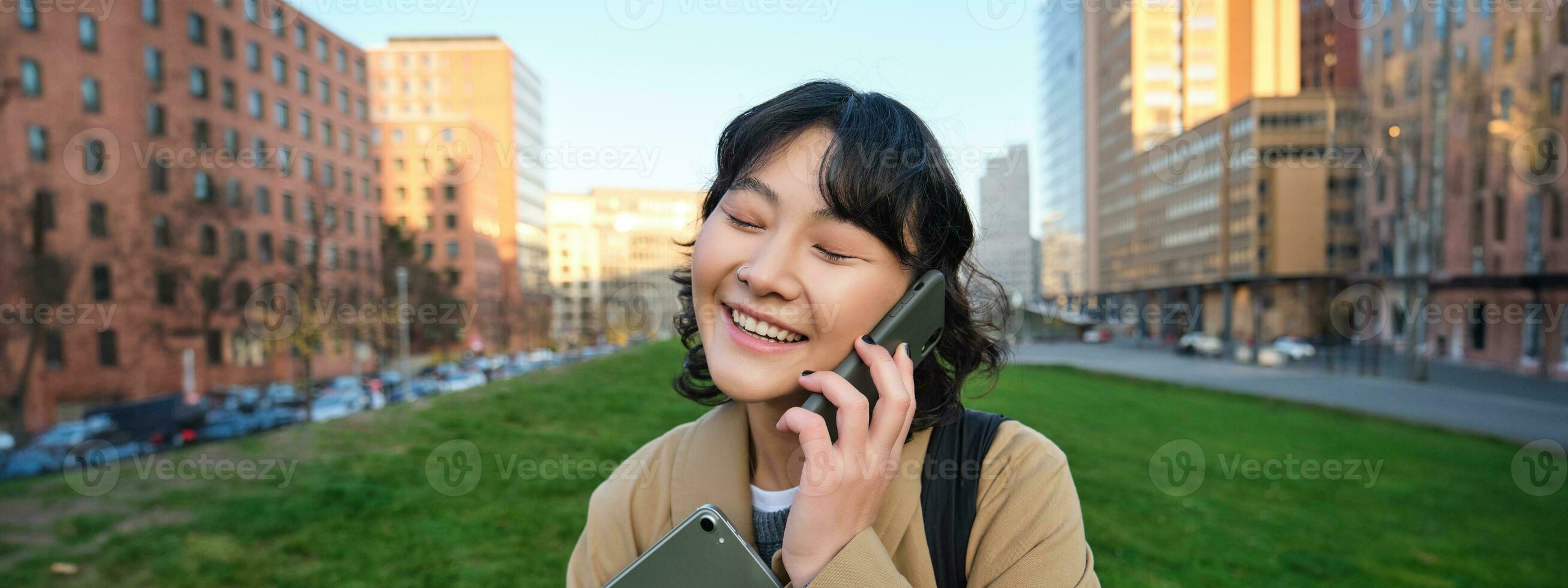 University lifestyle. Young asian student, girl with tablet and notebook, talks on mobile phone while walks from campus on street photo