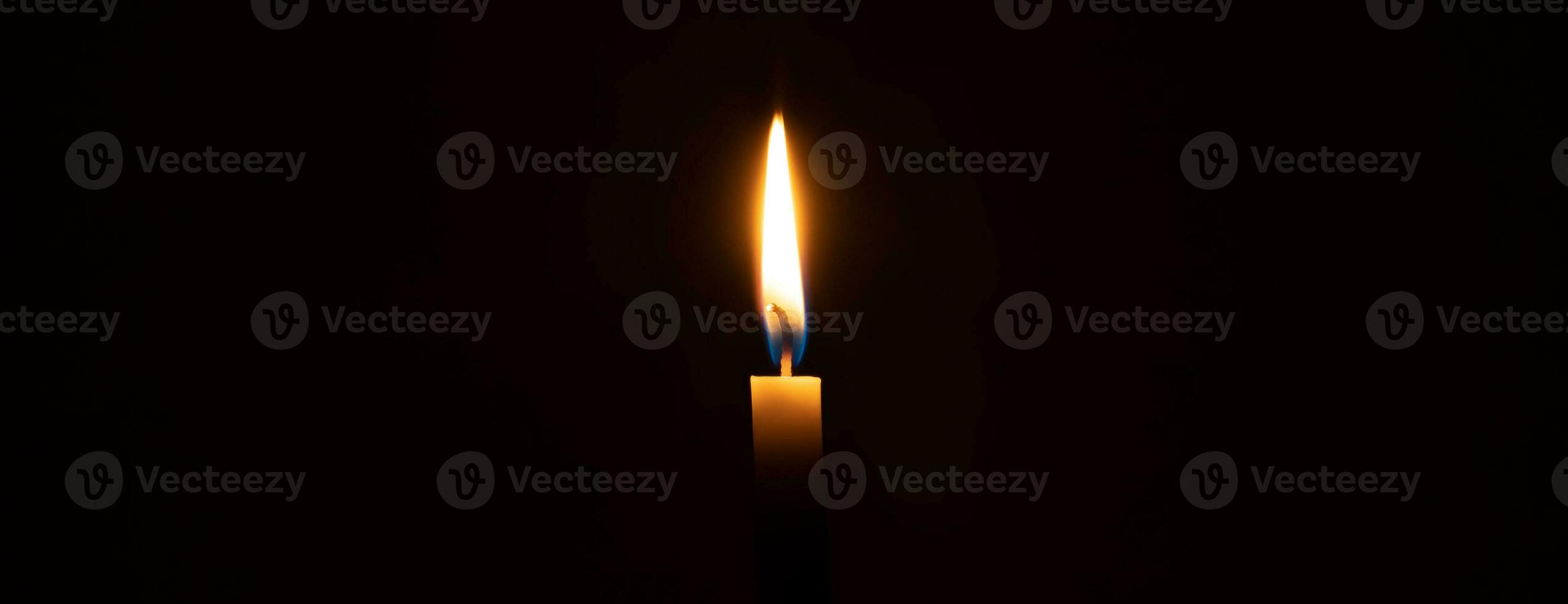 A single burning candle flame or light glowing on an orange candle on black or dark background on table in church for Christmas, funeral or memorial service with copy space photo