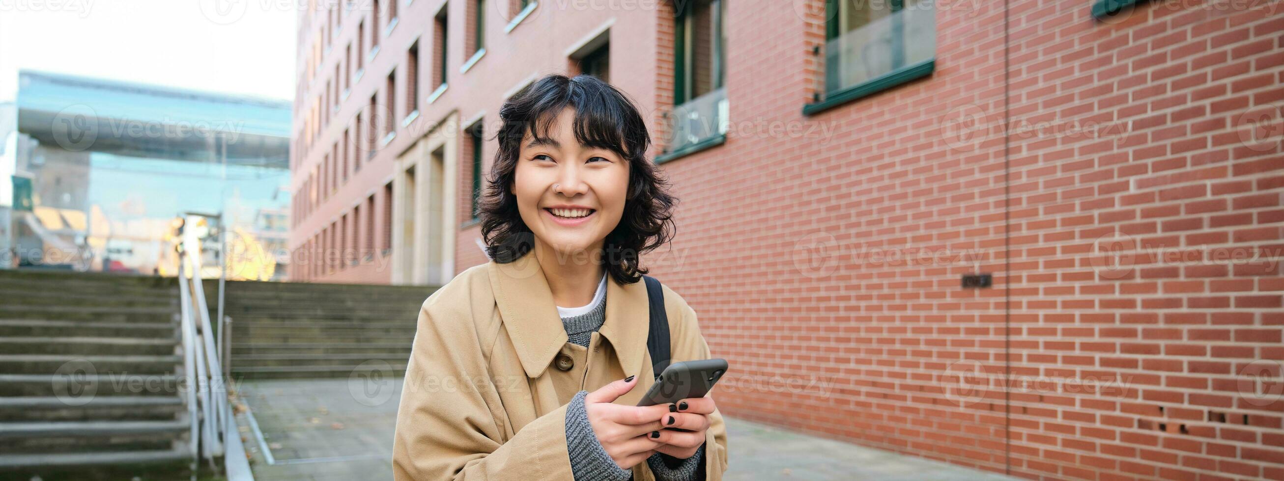 Stylish korean hipster girl, walks in city centre, sends text message, uses location map application on smartphone while walking down the street, wears trench coat photo