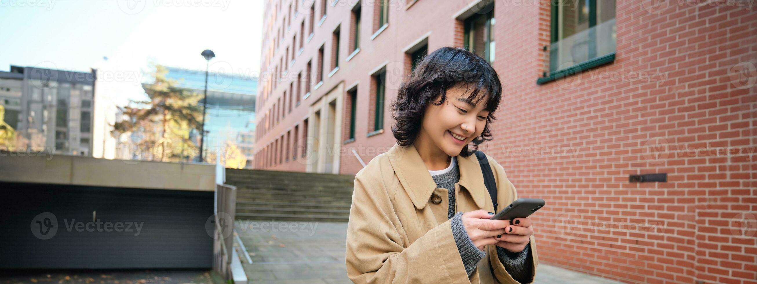 Stylish korean hipster girl, walks in city centre, sends text message, uses location map application on smartphone while walking down the street, wears trench coat photo