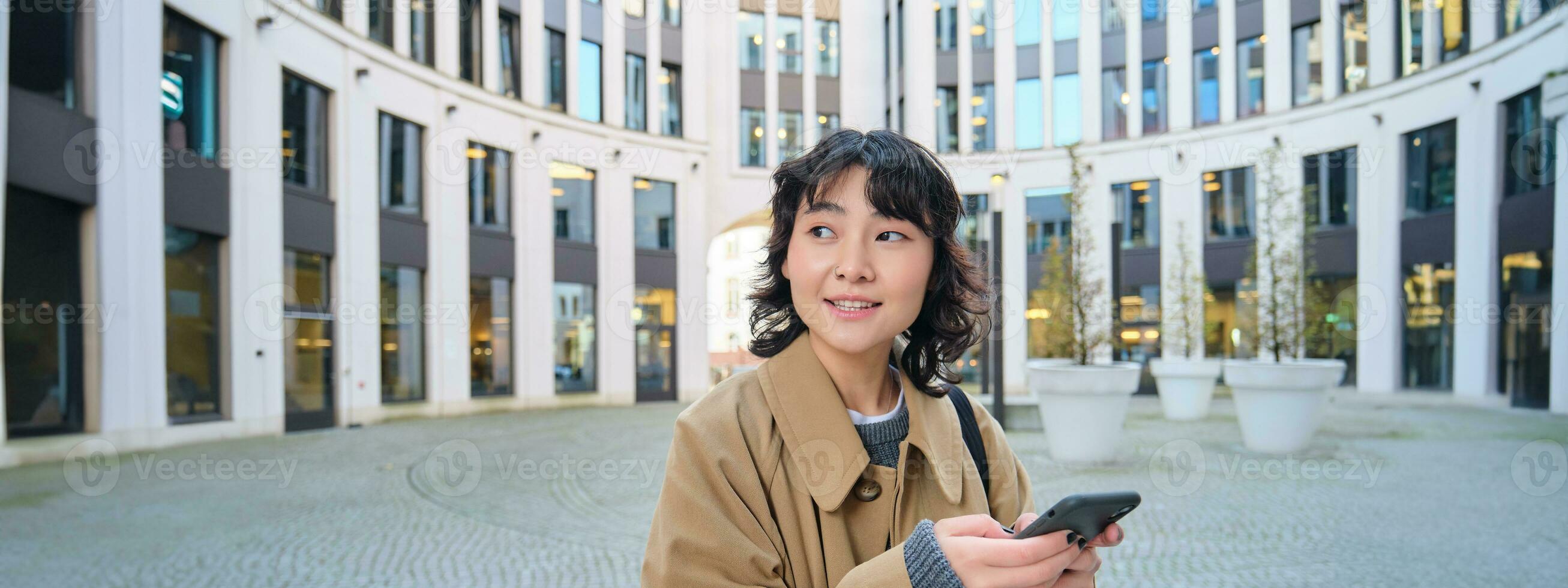 Stylish korean girl in headphones, listens music and uses mobile phone, stands in city centre, waits for someone on street and writes text message on her smartphone photo