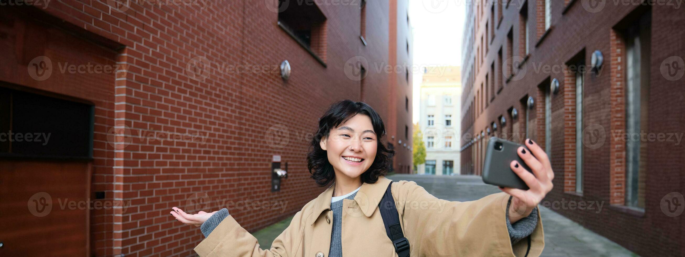 Happy korean girl, tourist takes photos on street, shows smth while records video of herself on smartphone, posing near building and smiling