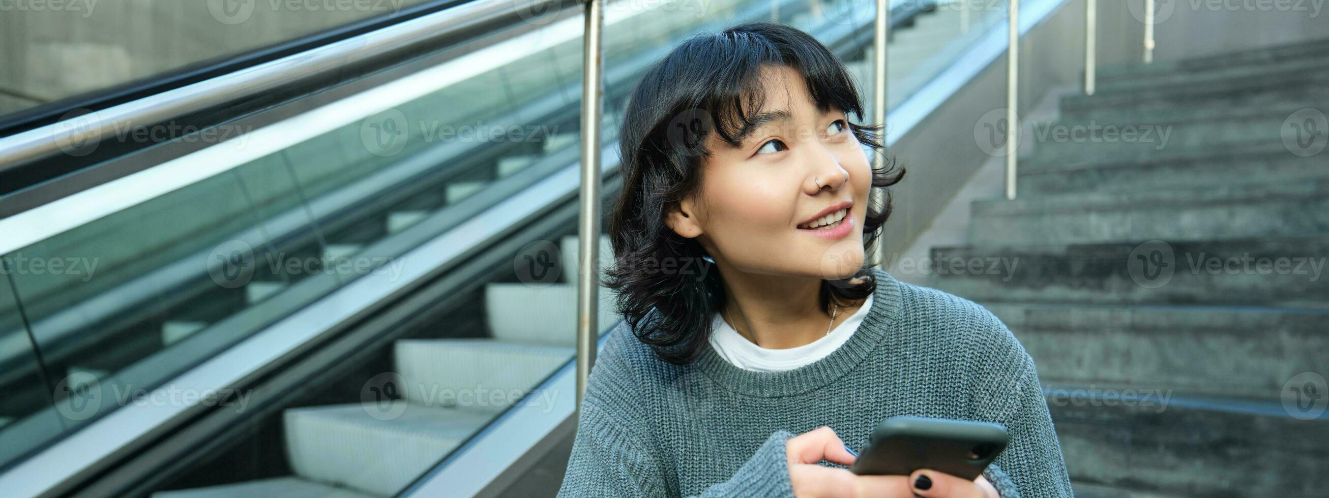 Stylish young girl student, sits on stairs with smartphone and backpack, laughs and smiles, texts message, uses social media photo