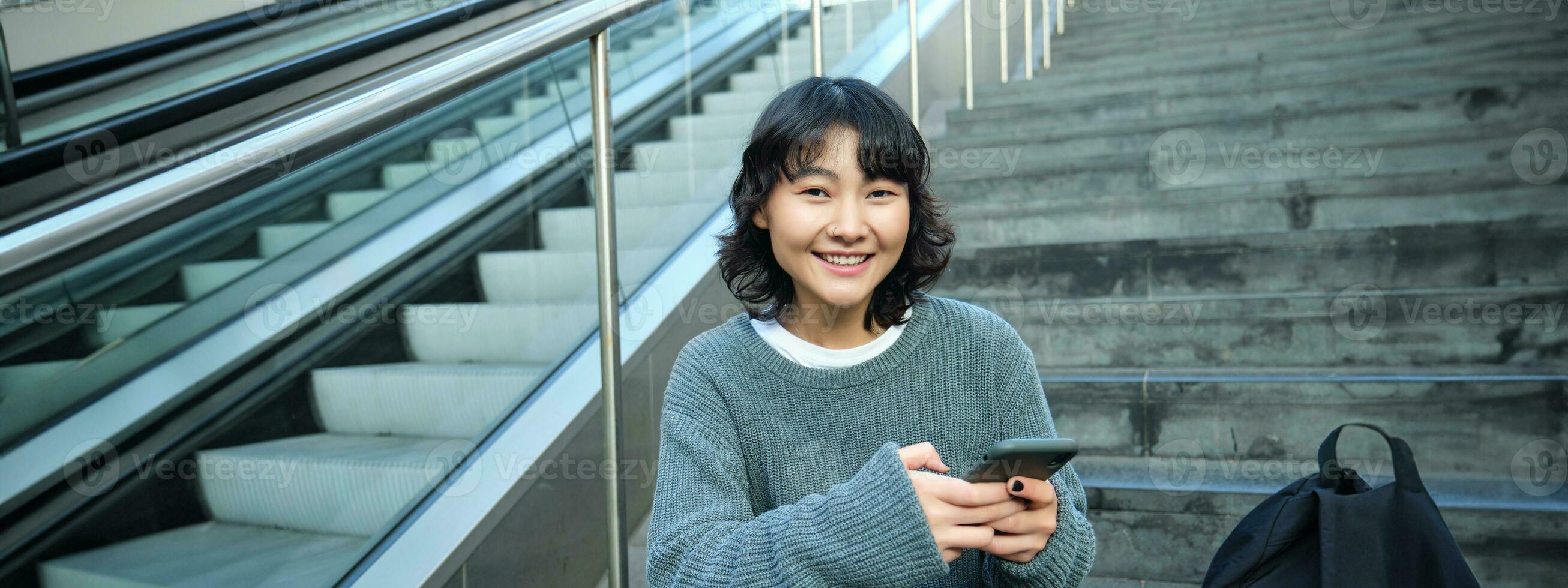 Stylish young girl student, sits on stairs with smartphone and backpack, laughs and smiles, texts message, uses social media photo
