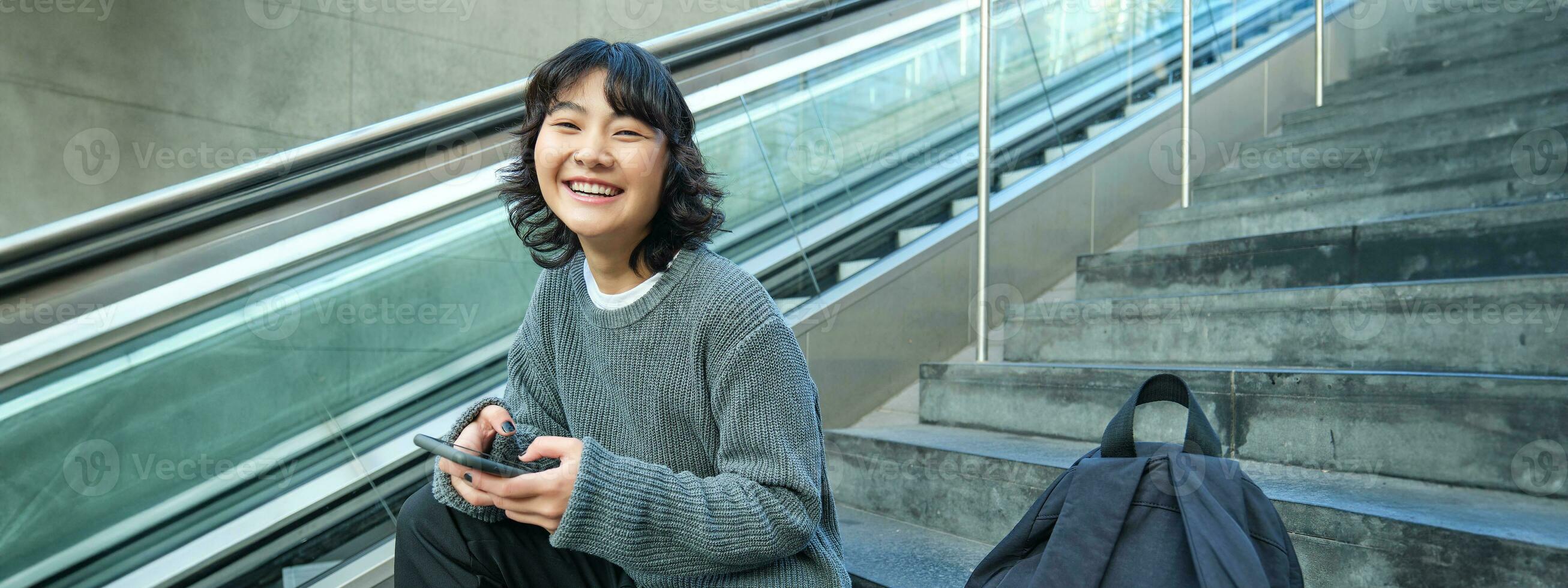 Stylish young girl student, sits on stairs with smartphone and backpack, laughs and smiles, texts message, uses social media photo