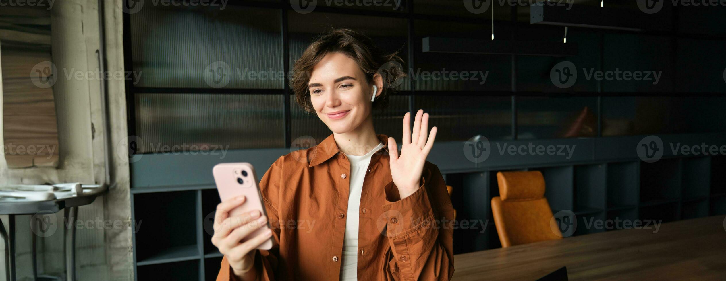 imagen de mujer conecta a trabajo llamar, vídeo charla en móvil teléfono, olas mano a teléfono inteligente aplicación y sonrisas, soportes en un oficina en casual ropa foto