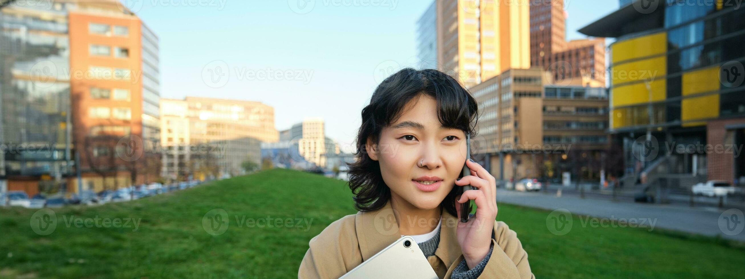 Stylish smiling girl, university student in trench coat, holds tablet, talks on mobile phone, has conversation over telephone and looks relaxed, stands on street photo