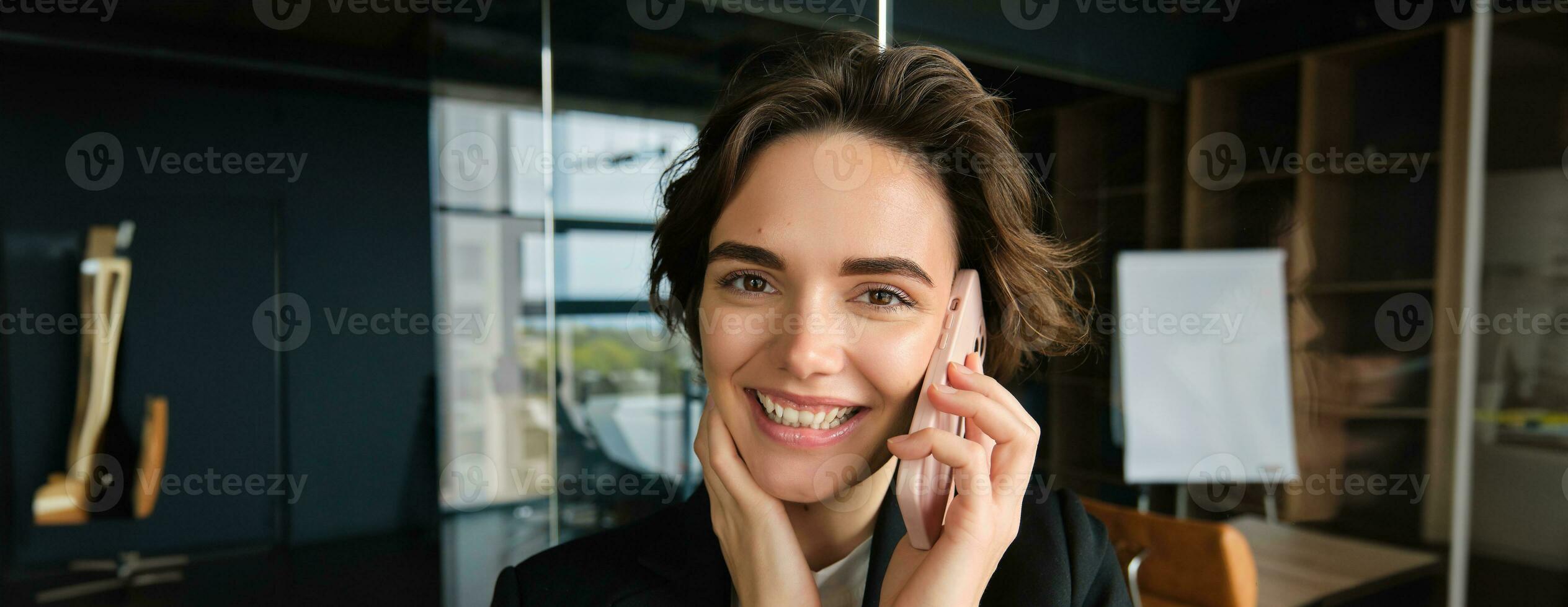 Close up portrait of smiling saleswoman, office manager stands in conference room, answers phone call, talking on mobile telephone with someone, receives great news and rejoices photo