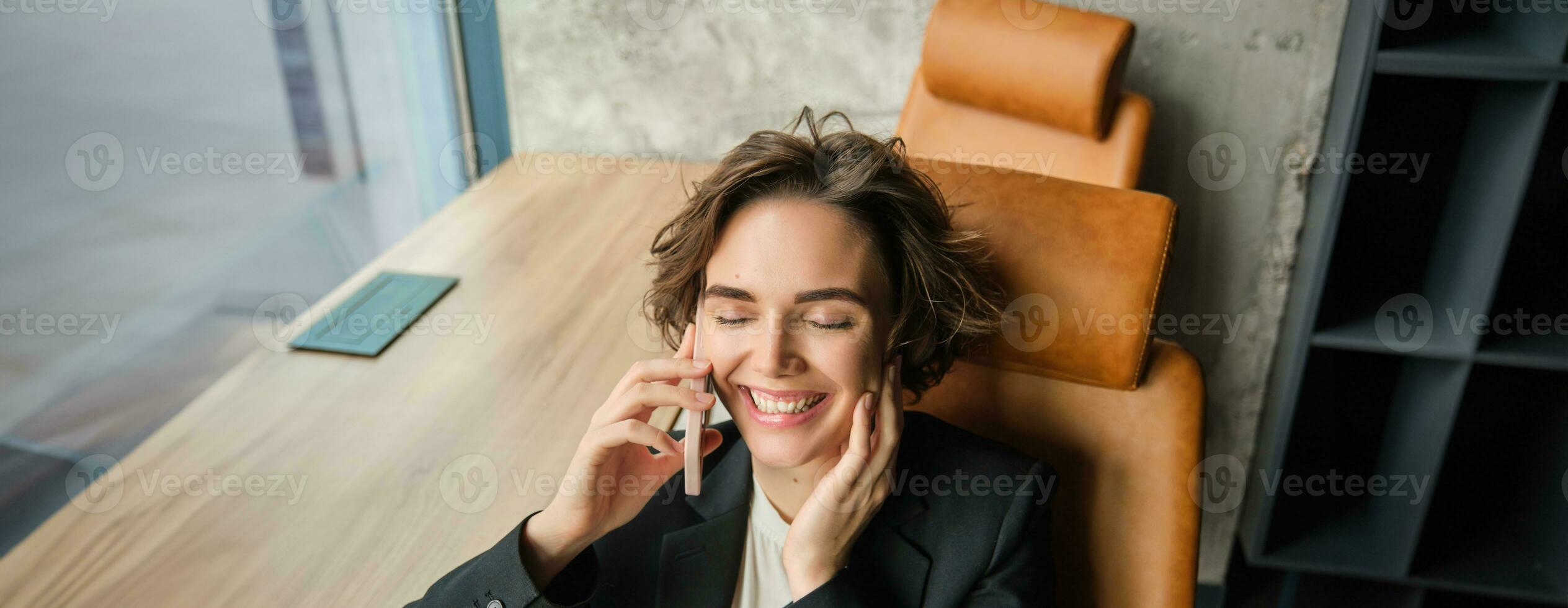Portrait of business woman in suit, sitting in her office and answering a phone call with pleased smile, having a negotiation over the telephone conversation photo