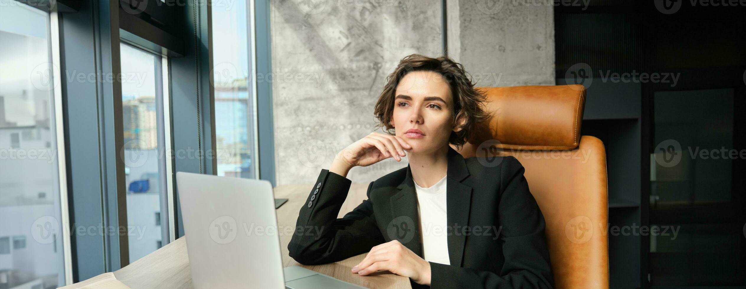 Portrait of confident businesswoman in office, sitting with laptop, looking serious outside window, thinking, solving issue at work photo