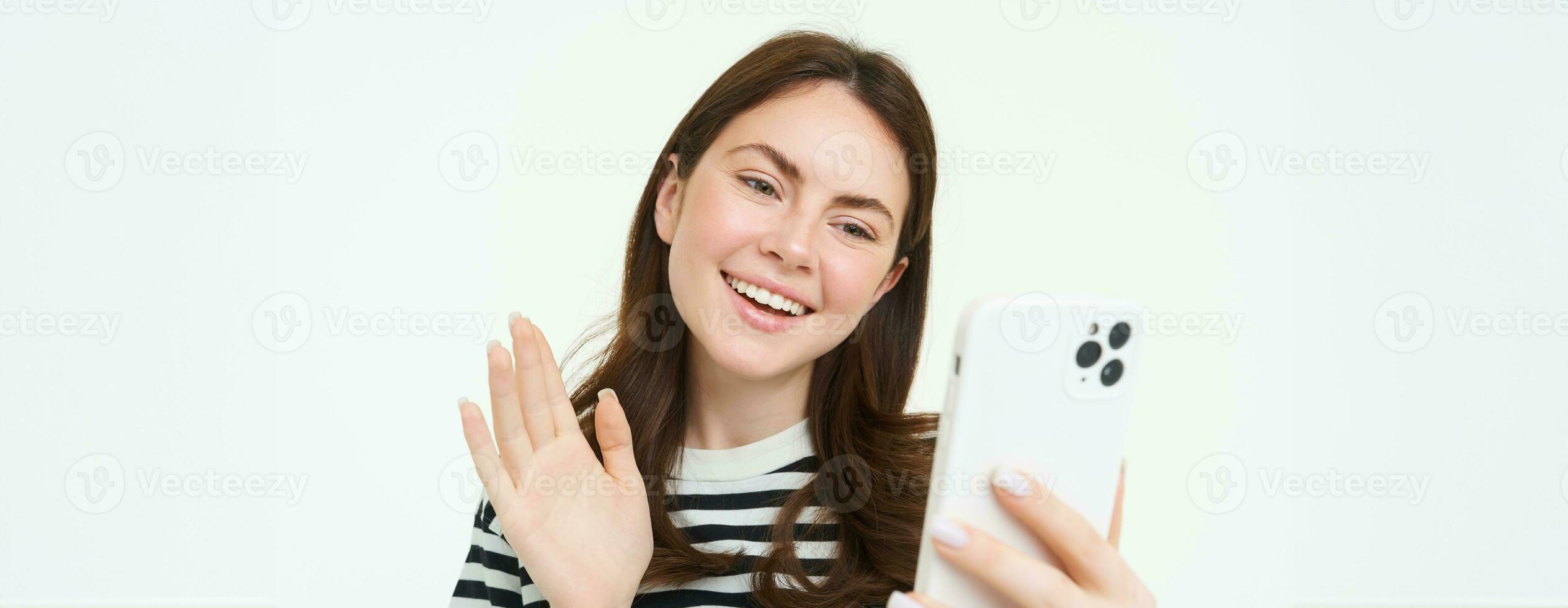 Happy young woman waves her hand at smartphone camera, saying hello to viewers on social media app, video chats on mobile phone app, white background photo