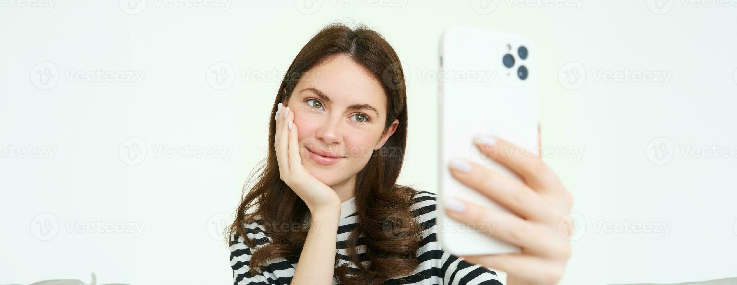 Portrait of young european woman taking selfie on smartphone, holding white mobile phone and posing for photos, isolated against white background photo