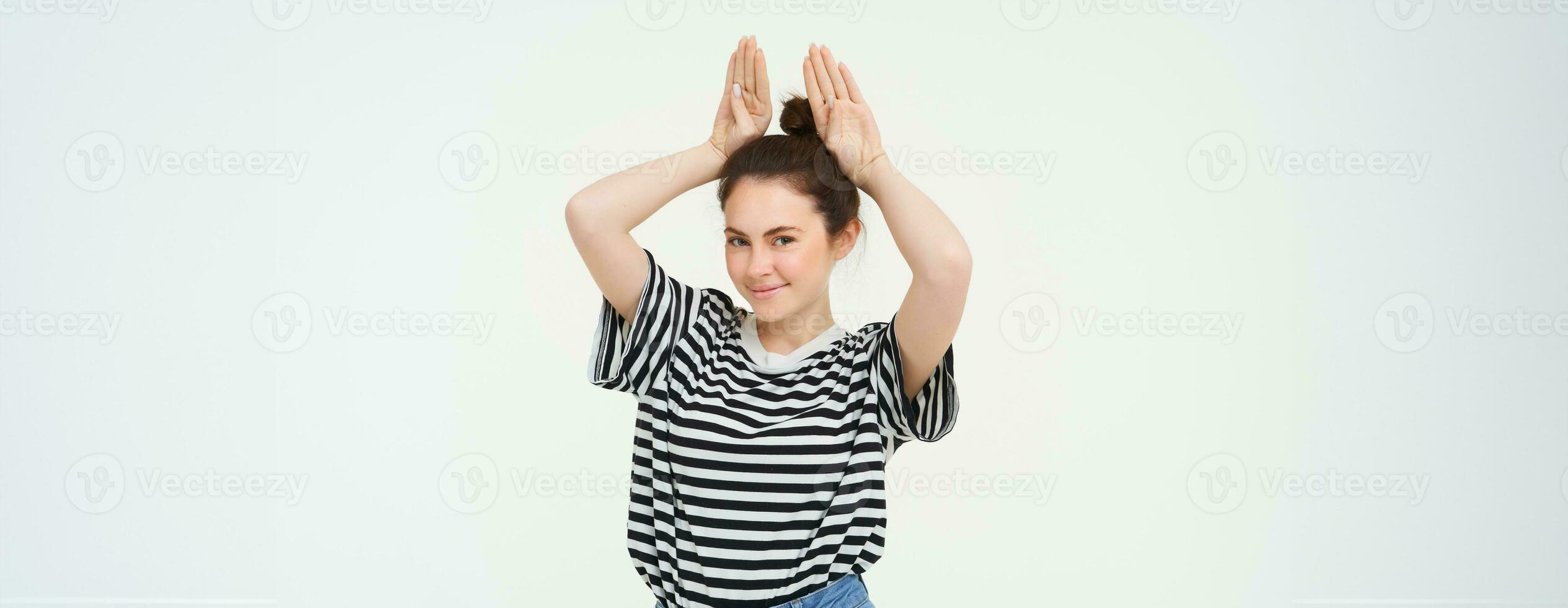 Portrait of cute caucasian woman, makes animal ears, cute pose, smiling at camera, standing over white background photo