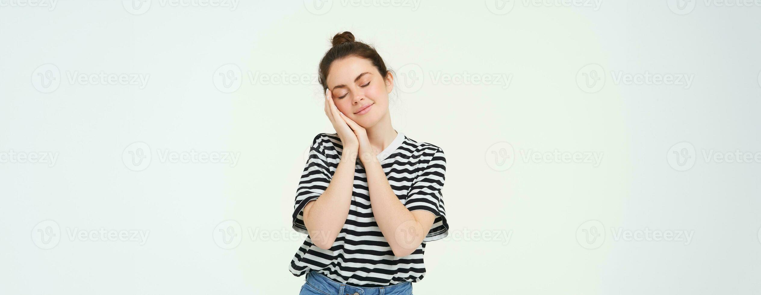 Image of tender woman, 25 years, holds hands near face and closes eyes, sleeps, smiles pleased during nap, isolated against white background photo
