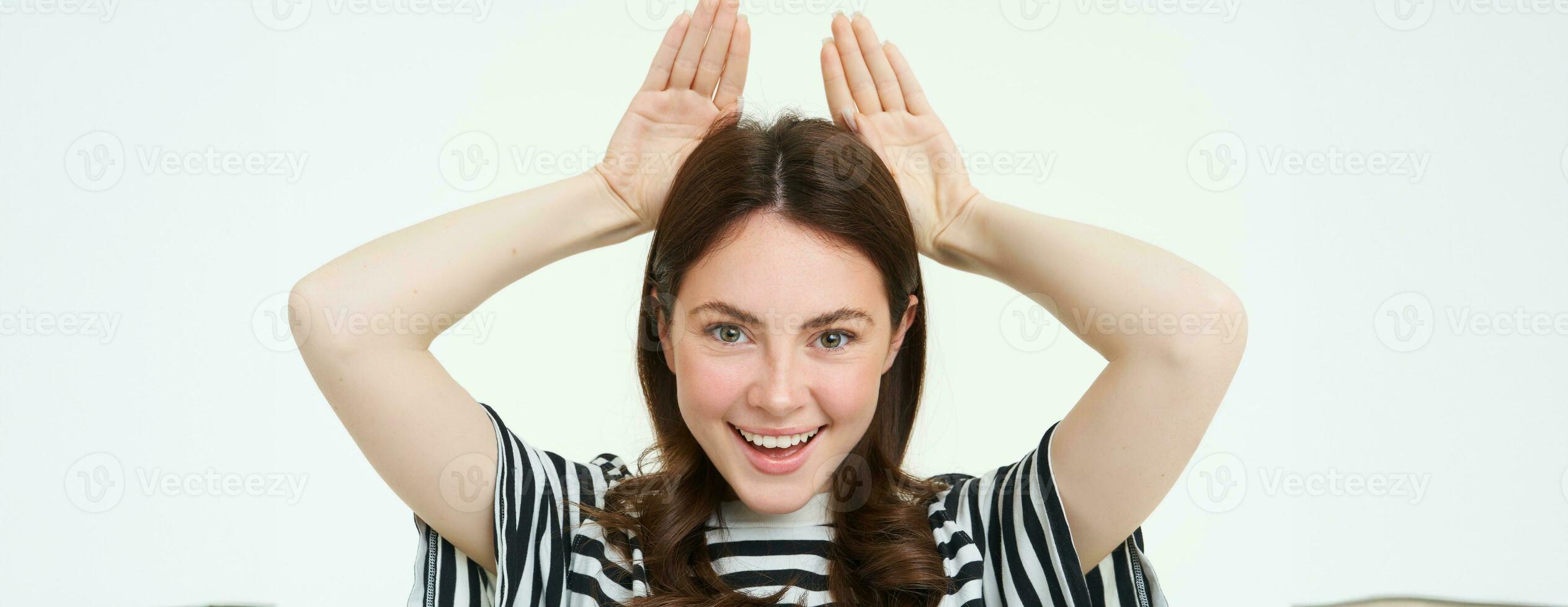 Image of cute brunette girl shows animal eats with hands on top of head, smiling and looking happy, white background photo