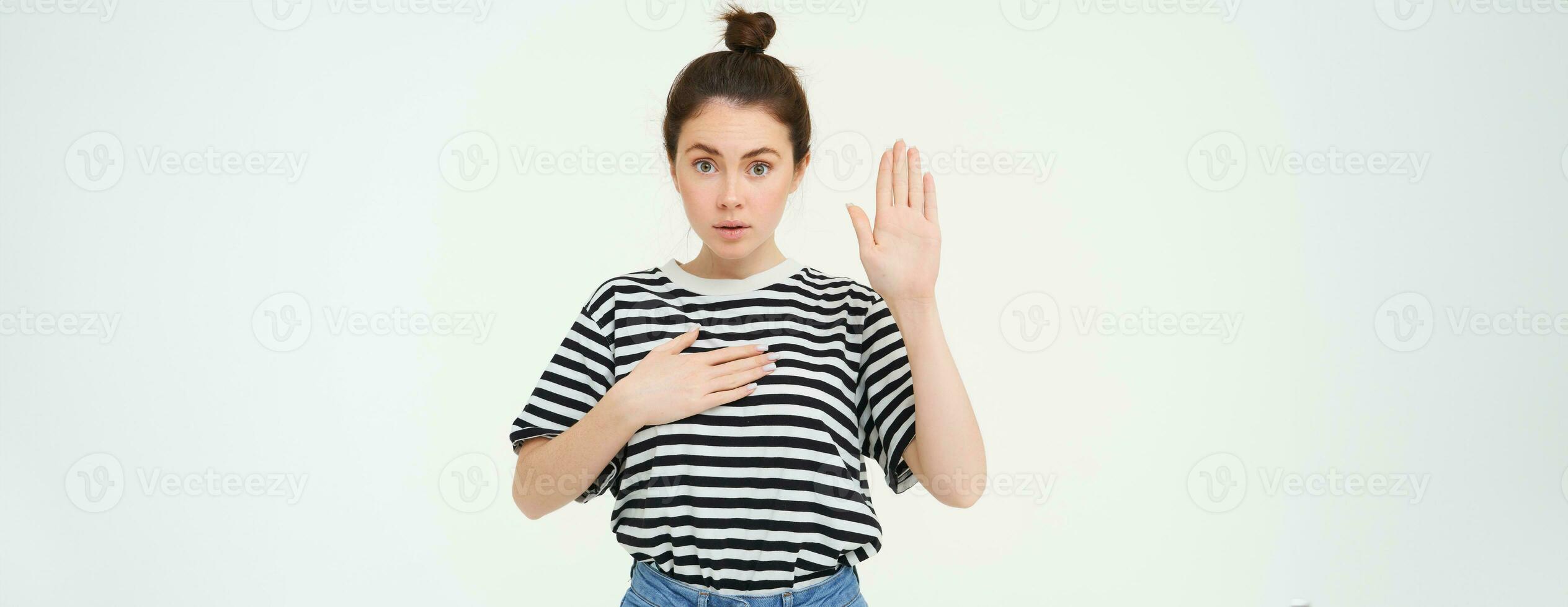 Image of woman makes promise, swears, raises one hand and puts palm on heart, being honest, stands over white background photo