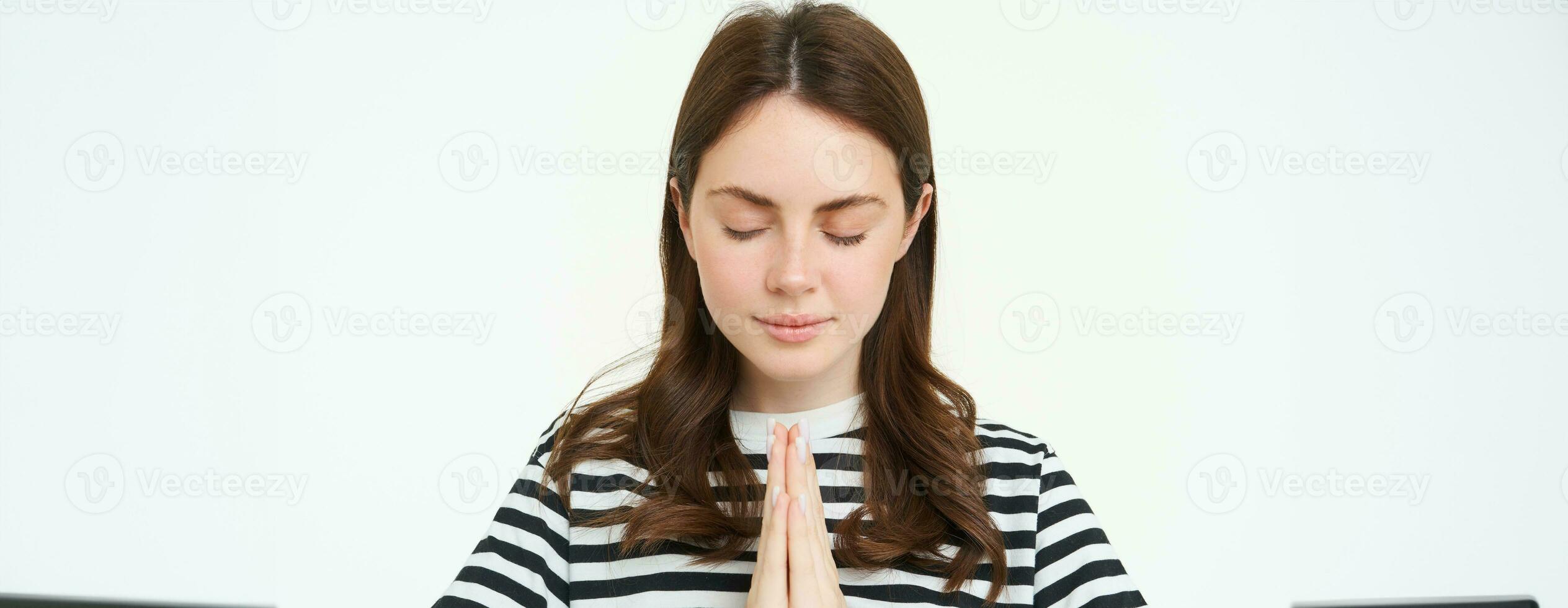 Portrait of woman showing namaste gesture, holding palms clenched together near chest, standing isolated over white background photo