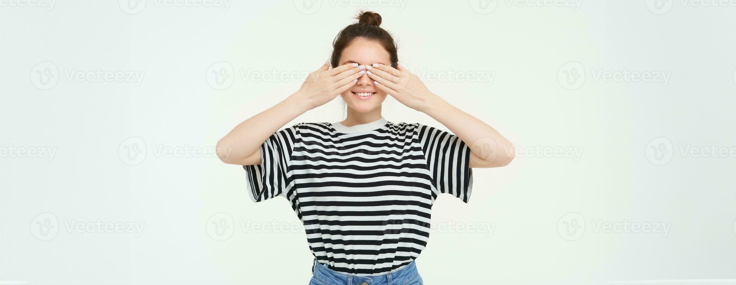 Portrait of girl waiting for surprise, covers eyes with hands, peeking through fingers, stands over white background photo