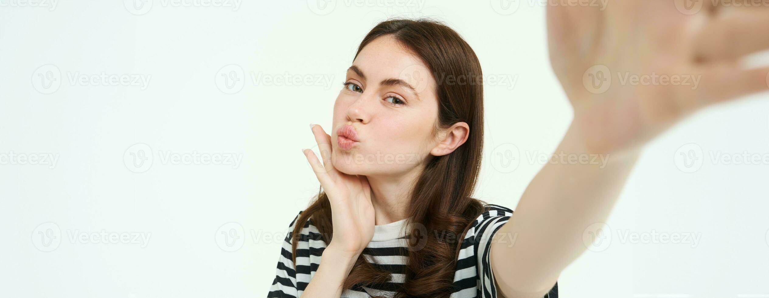 Portrait of cute, feminine girl posing near something, taking selfie on smartphone, holding camera with extended hand, isolated on white background photo