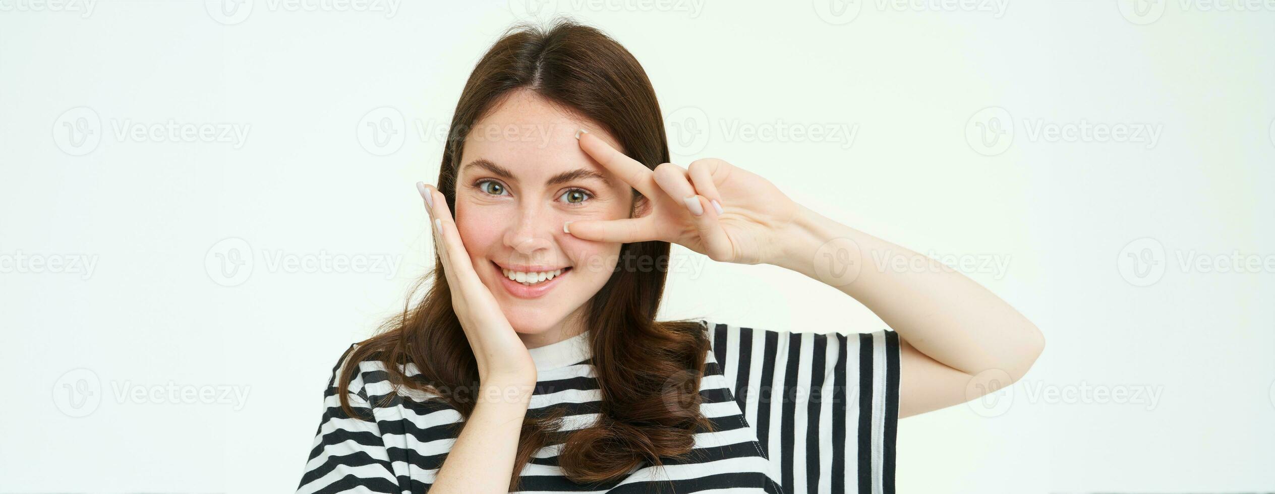 Portrait of beautiful, smiling female model, touches her clear glowing skin, shows peace sign and looks happy at camera, isolate on white background photo