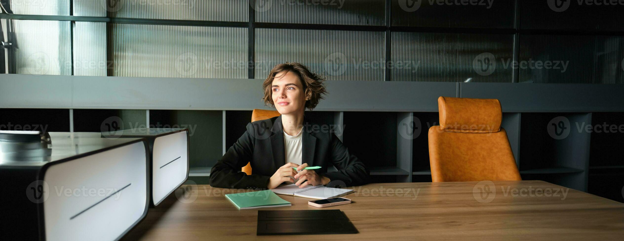 Young businesswoman waiting for start of meeting. Corporate woman in suit sitting in office, working on documents, waiting for client photo