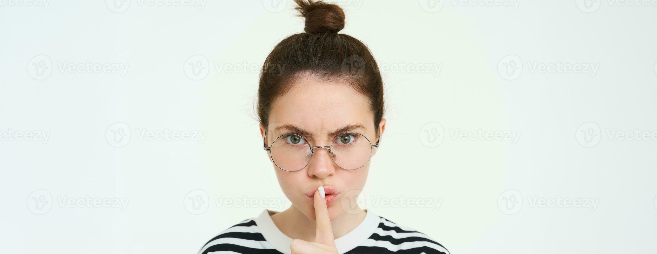 Image of serious woman in glasses, teacher shushing, makes shush, hush gesture, press finger to lips and frowning from disapproval, standing over white background photo