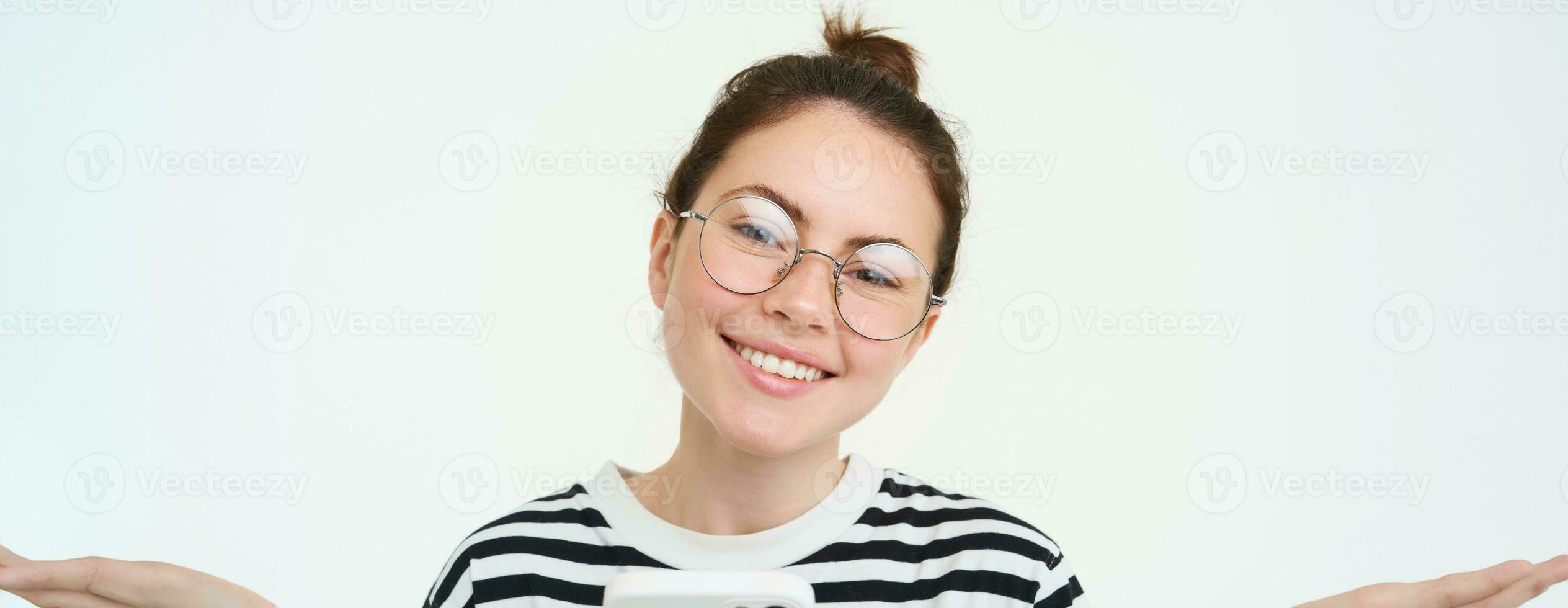 imagen de joven mujer en anteojos, utilizando su móvil teléfono, en pie con teléfono inteligente y sonriente, en pie terminado blanco antecedentes foto