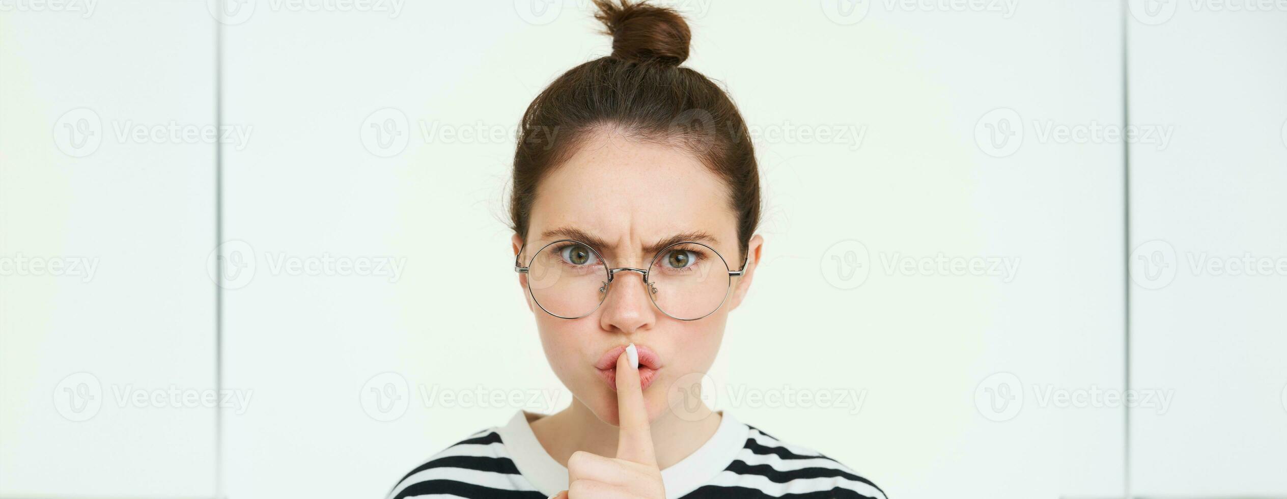 Image of serious woman in glasses, teacher shushing, makes shush, hush gesture, press finger to lips and frowning from disapproval, standing over white background photo