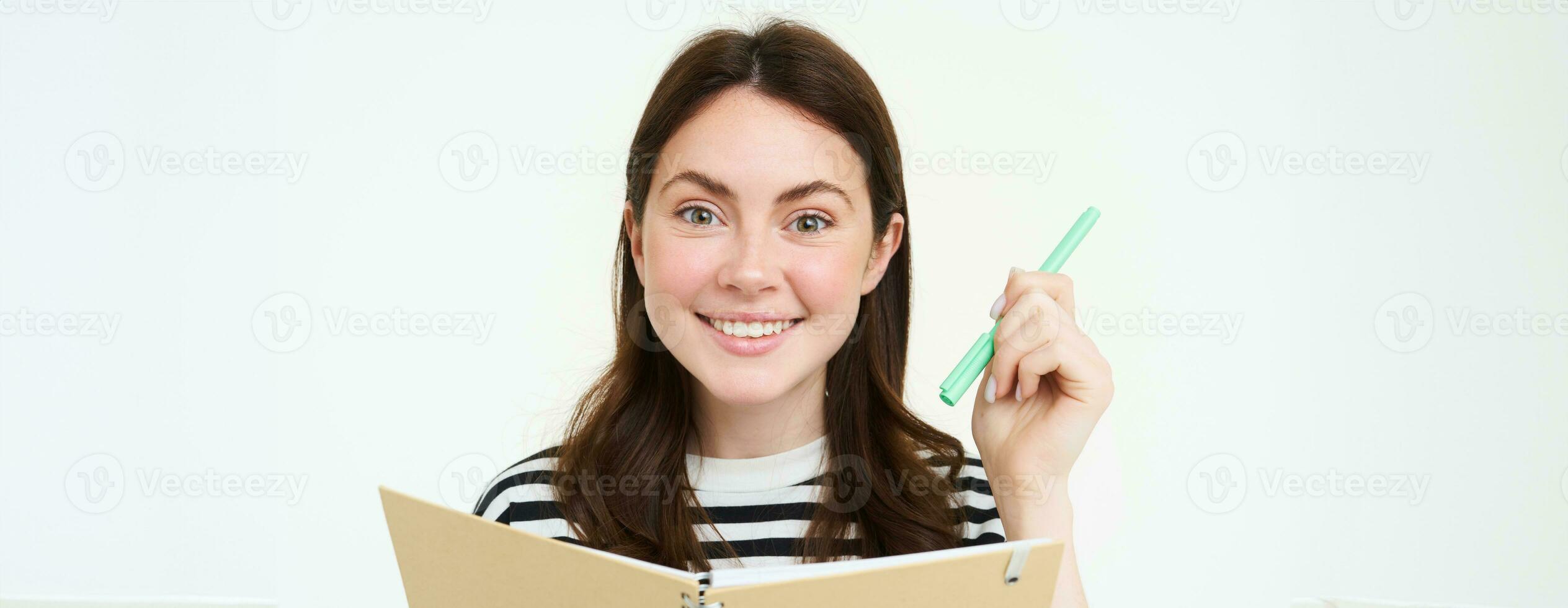 Portrait of creative smiling girl, writing down memos in planner, holding pen and notebook, makes notes, stands over white background photo