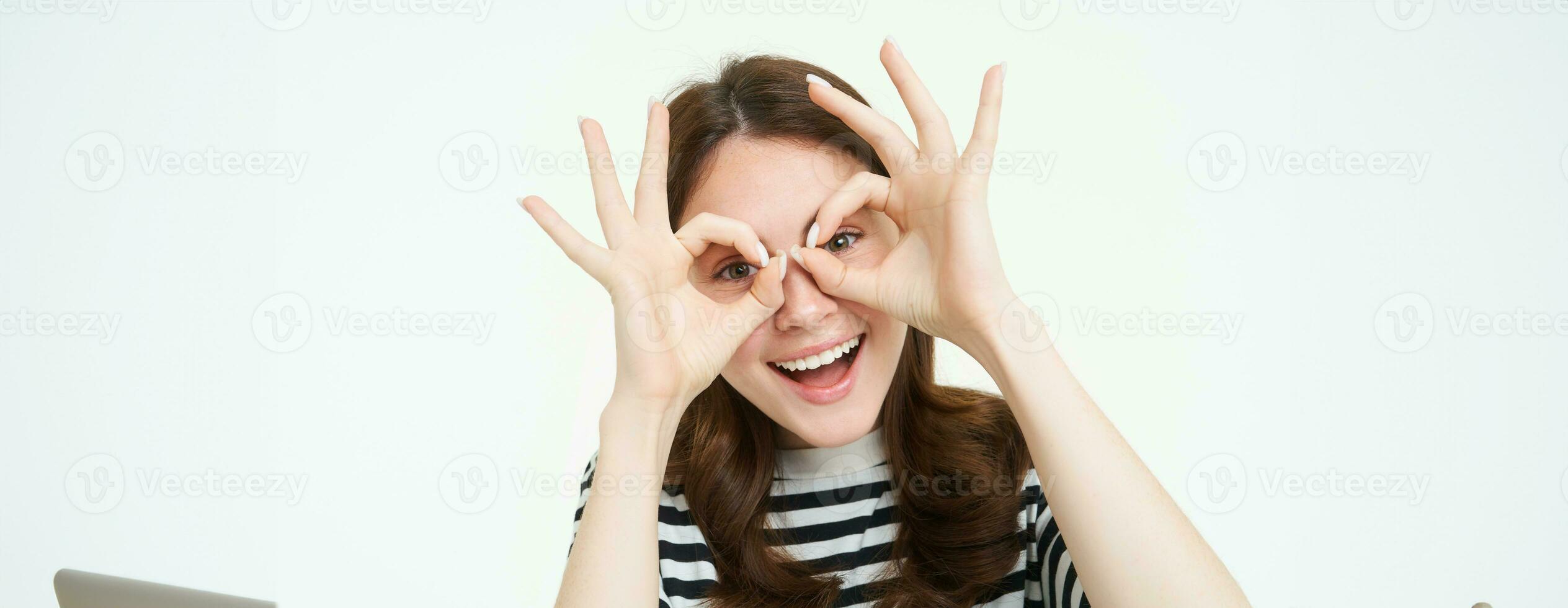 Portrait of happy, beautiful young woman, makes glasses with fingers around eyes and laughing, having fun, enjoying event, standing over white background photo