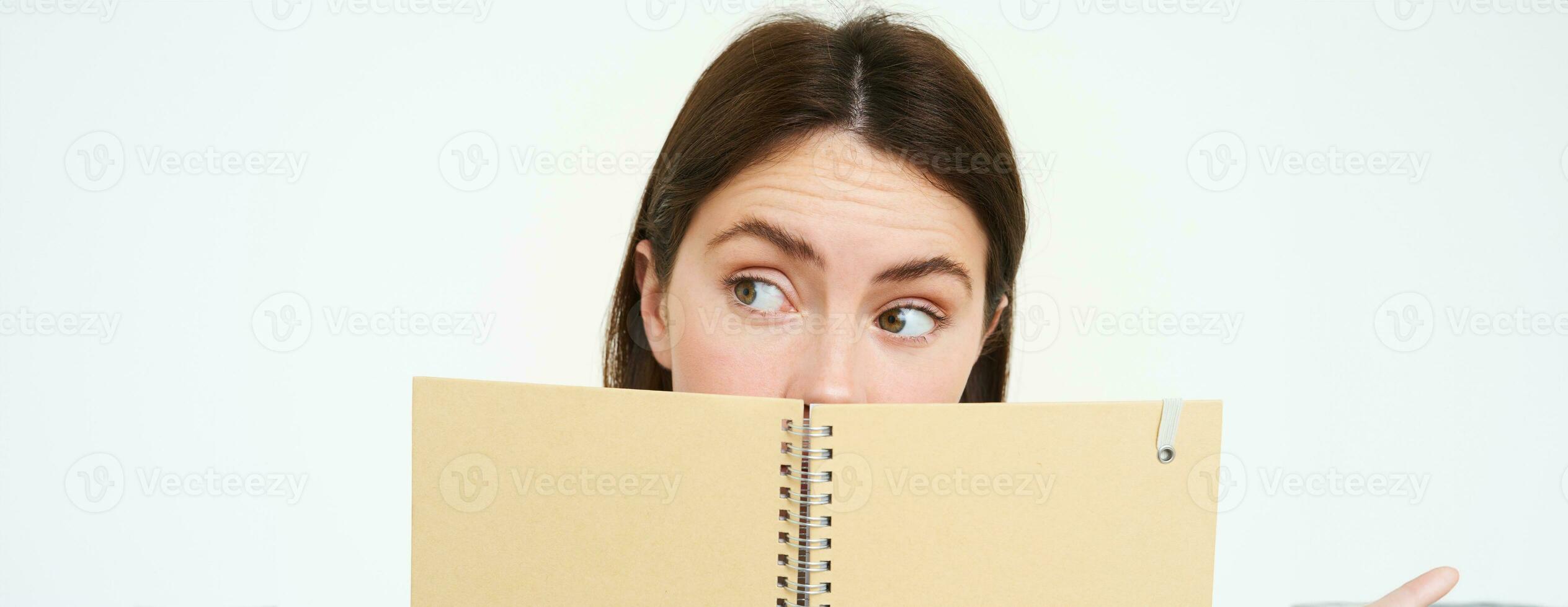 Close up portrait of woman with thoughtful expression, looking aside, hiding half of face behind notebook, standing with memo planner over white background photo