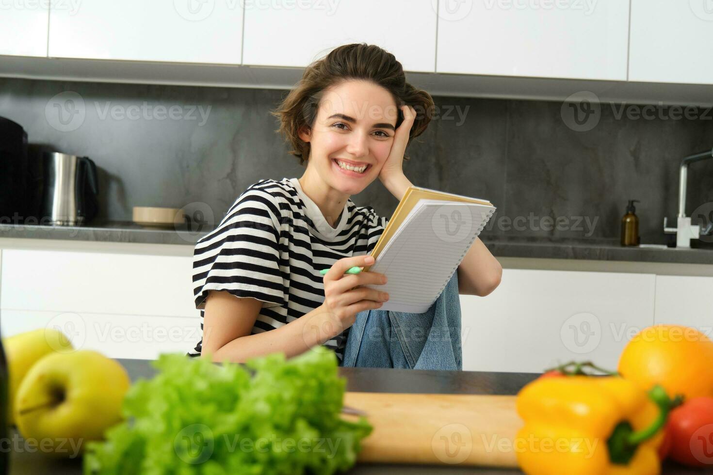 Portrait of young smiling woman in kitchen, holding notebook, making notes for recipe, writing grocery list, cooking salad, sitting near vegetables and chopping board photo