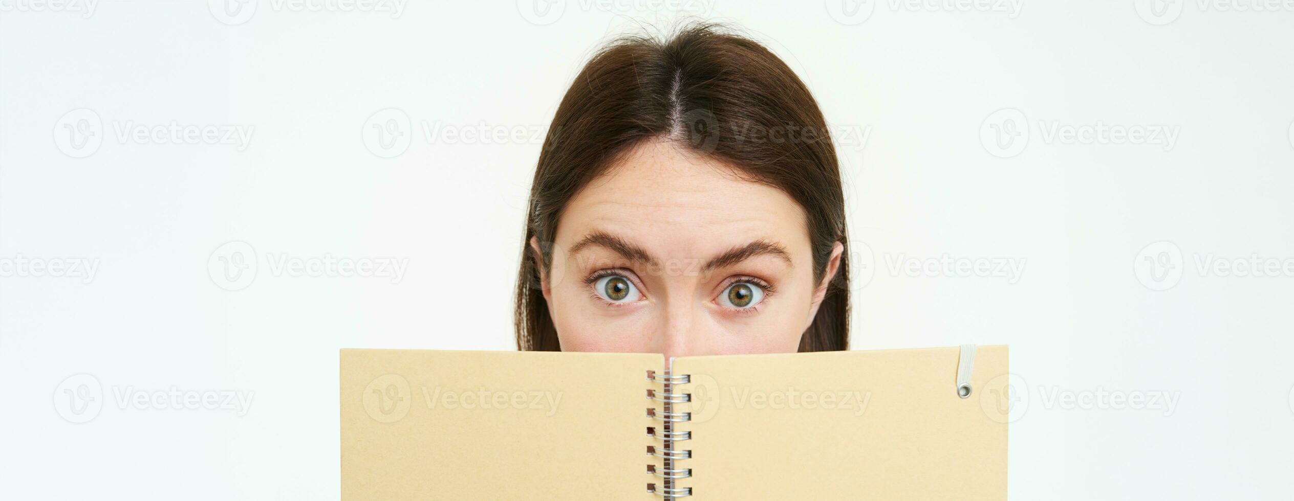 Close up portrait, half face of woman looking surprised, raising her eyebrows and staring at camera, holding notebook, daily planner organizer, isolated against white background photo
