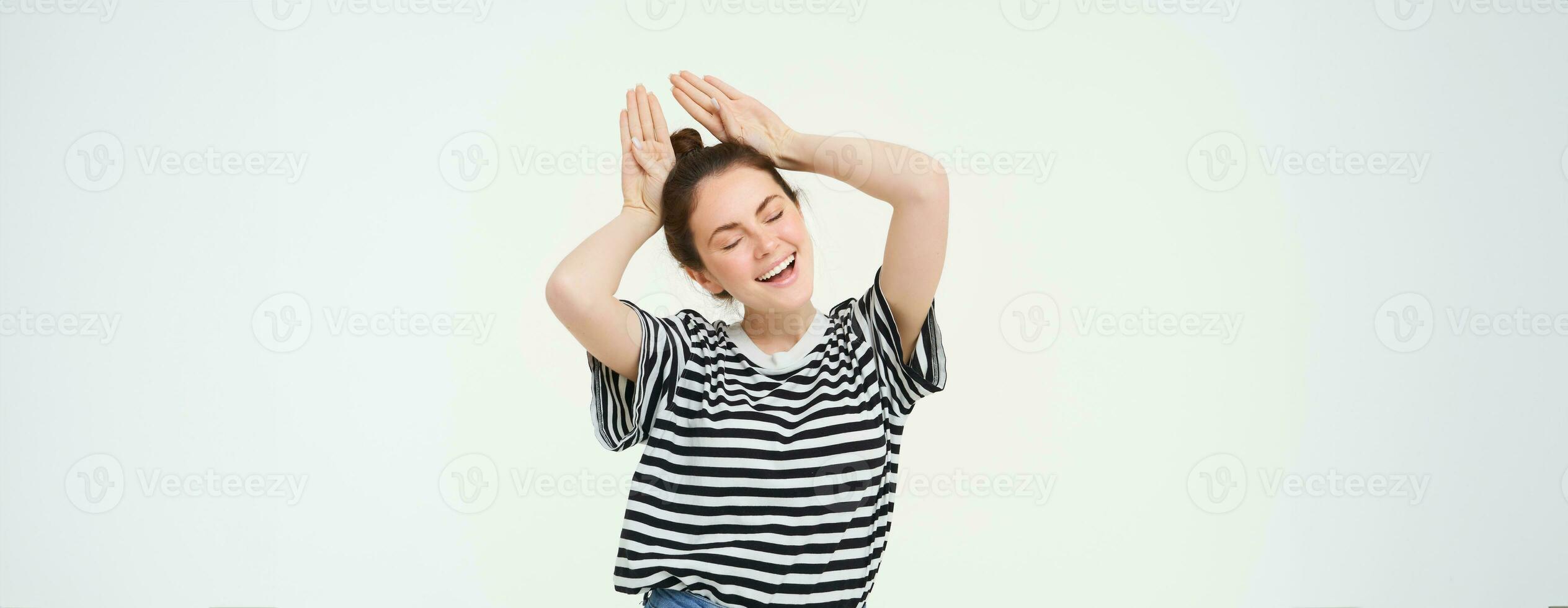 Portrait of cute caucasian woman, makes animal ears, cute pose, smiling at camera, standing over white background photo