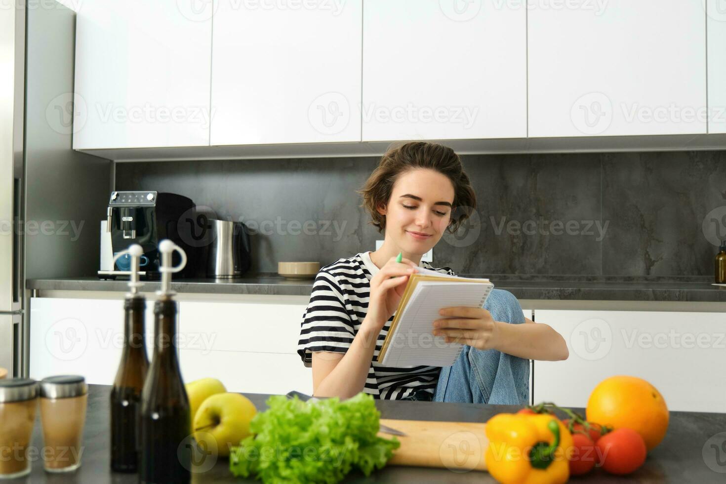 Portrait of young smiling woman in kitchen, holding notebook, making notes for recipe, writing grocery list, cooking salad, sitting near vegetables and chopping board photo