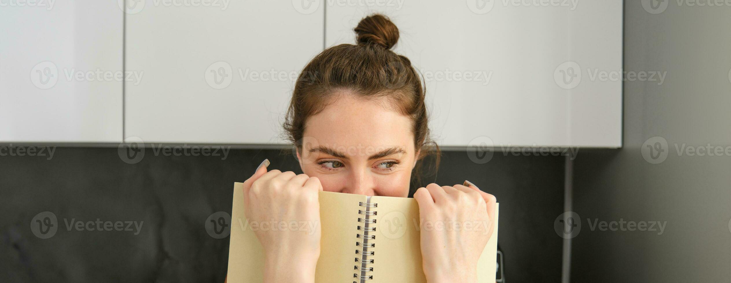 Cute woman hides face behind notebook, looking aside, smiling with eyes photo