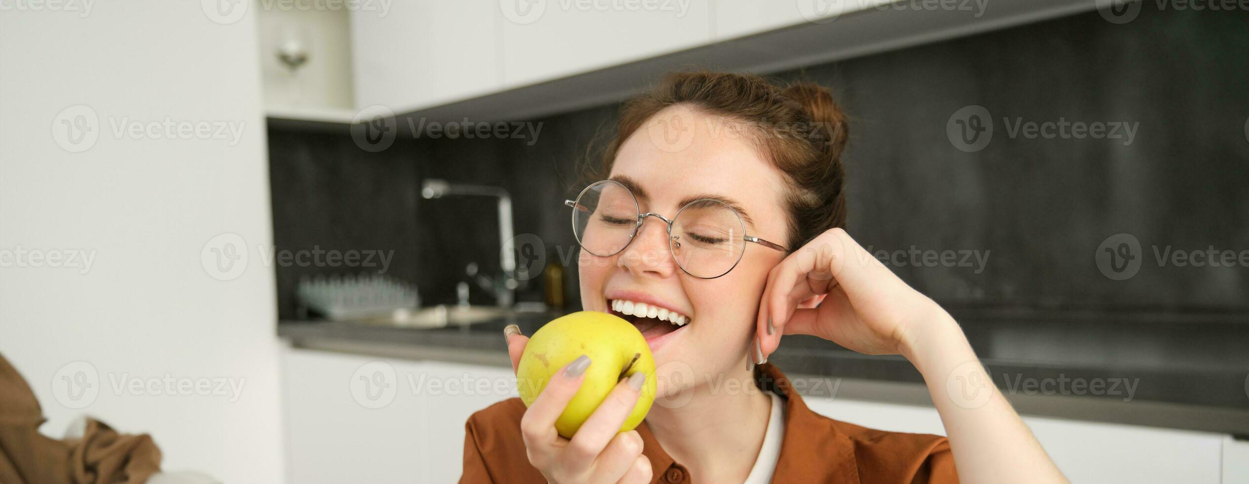 Portrait of smiling, modern woman in glasses, sitting at home and eating green apple, having a lunch break, biting fruit photo