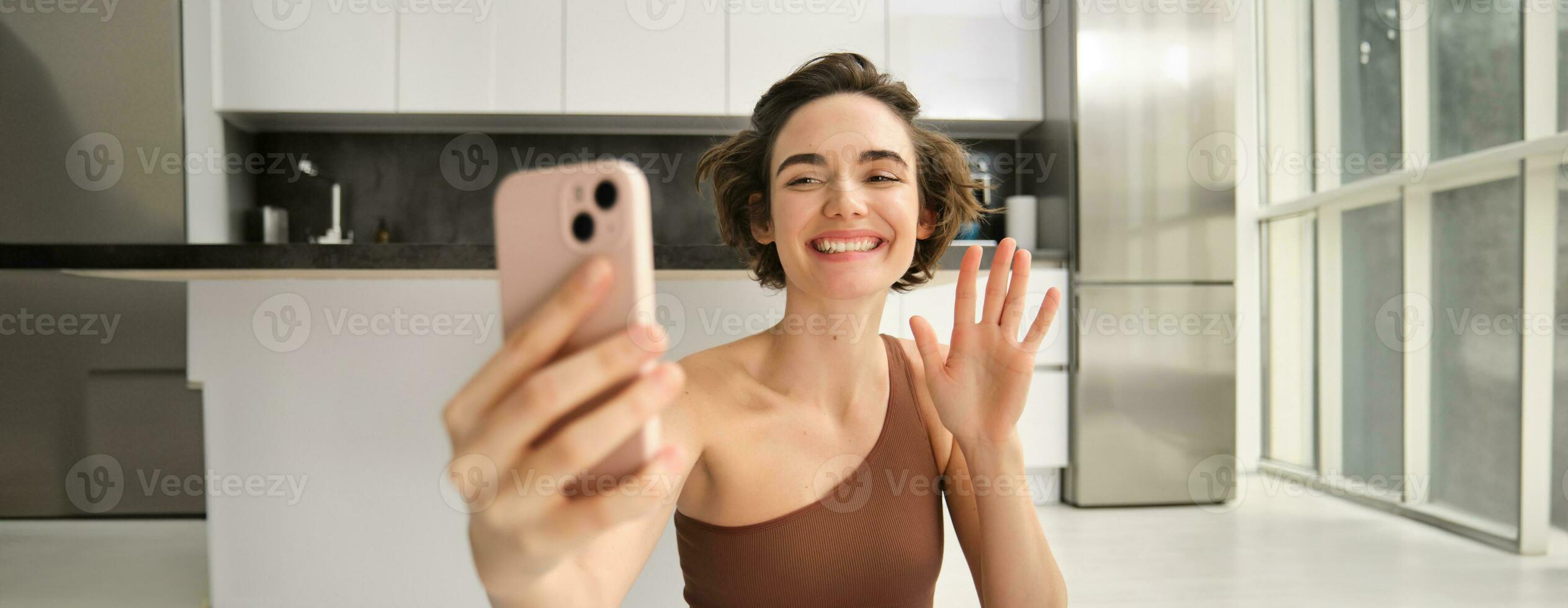 hermosa joven mujer utilizando móvil teléfono y rutina de ejercicio a hogar, ondulación mano a teléfono inteligente, vídeo charlas, haciendo yoga formación En Vivo en línea, registros Deportes vlog en su casa foto