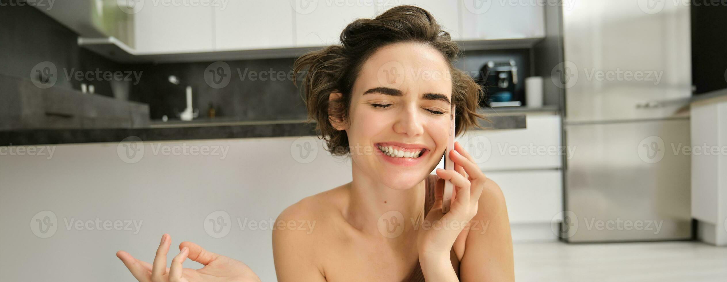 Close up portrait of happy smiling woman talking on mobile phone from her kitchen. Chatty girl with smartphone, laughing, calling someone photo