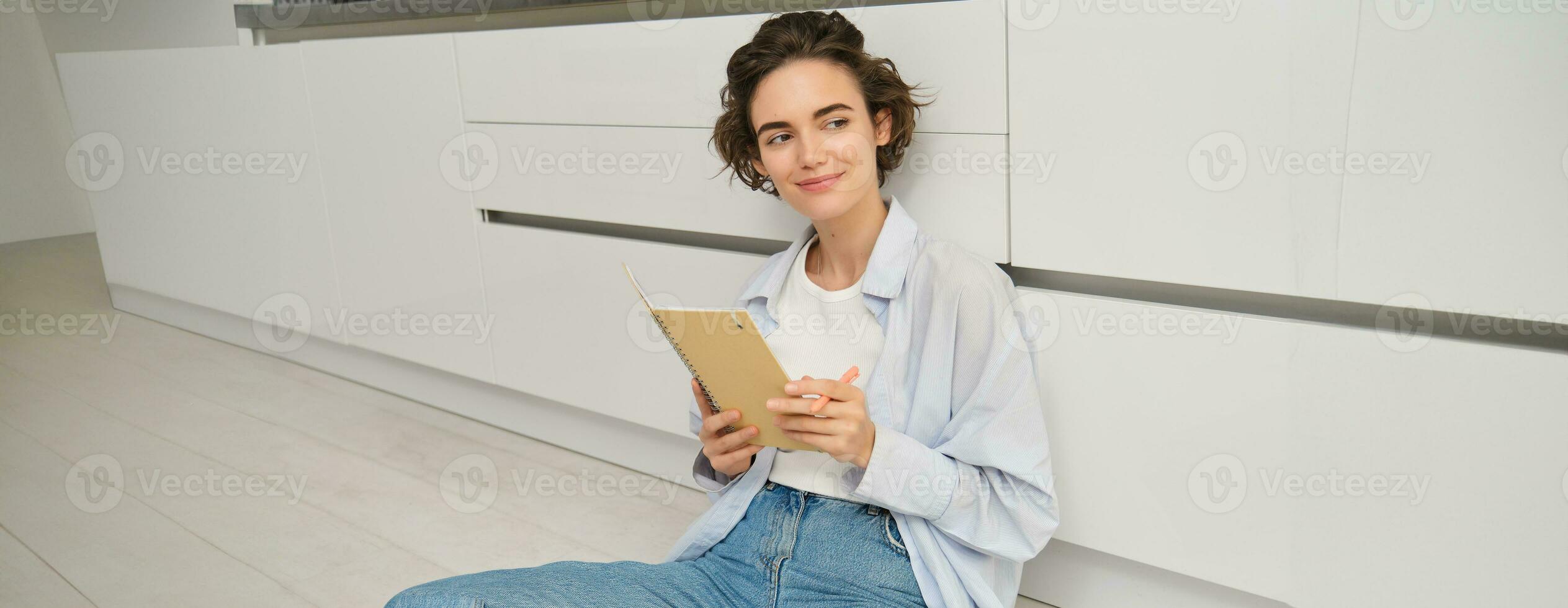 Portrait of creative girl, draws sketches, writes in diary, holds her notebook journal and sits on floor in kitchen, smiles and looks outside the window photo