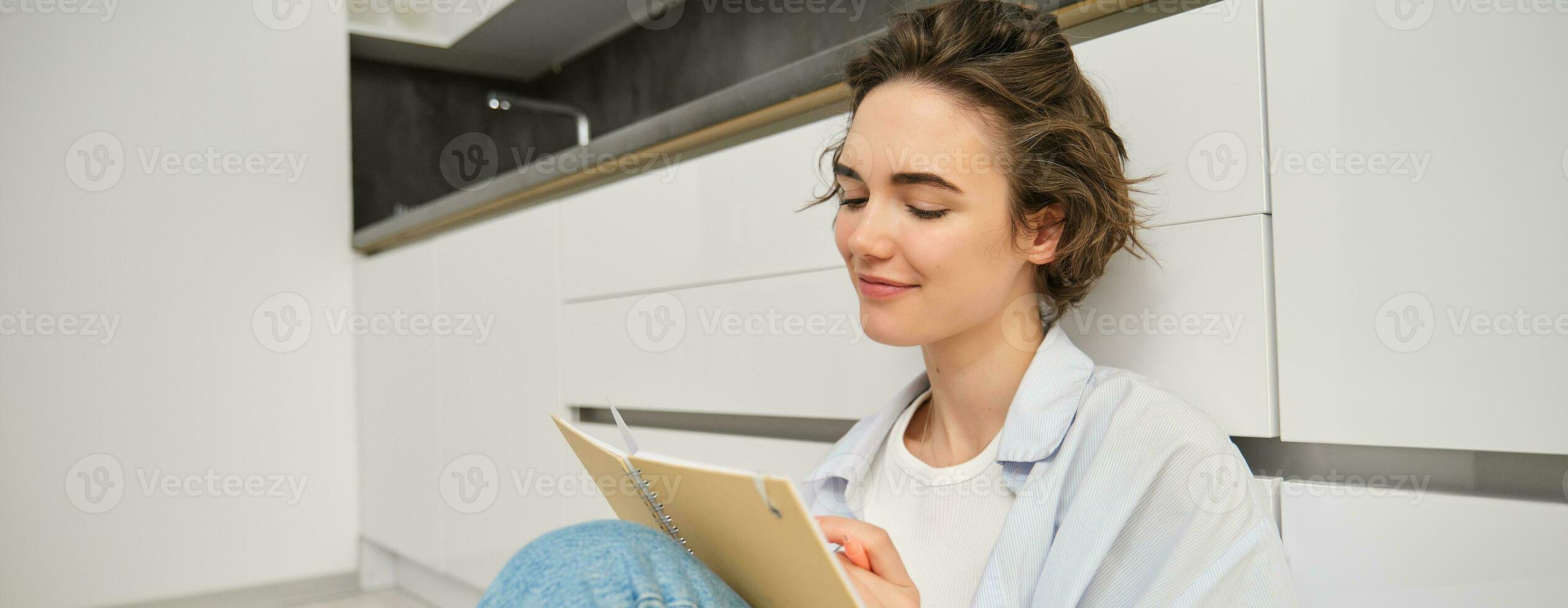Close up portrait of creative, smiling young woman, sits on floor with pen and paper, writes in journal, makes notes, does homework, puts notes in her planner photo