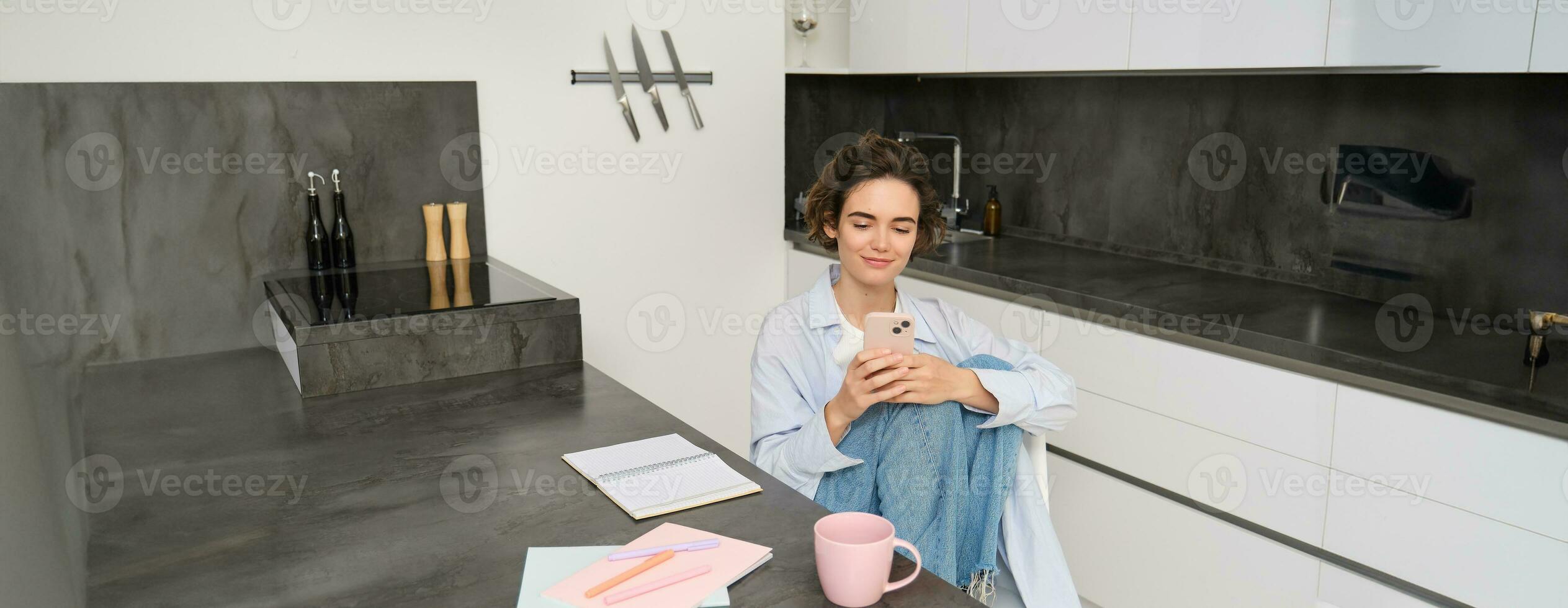 Young woman having break from work, sitting in kitchen and using smartphone, girl studies at home, sends a message on mobile phone photo