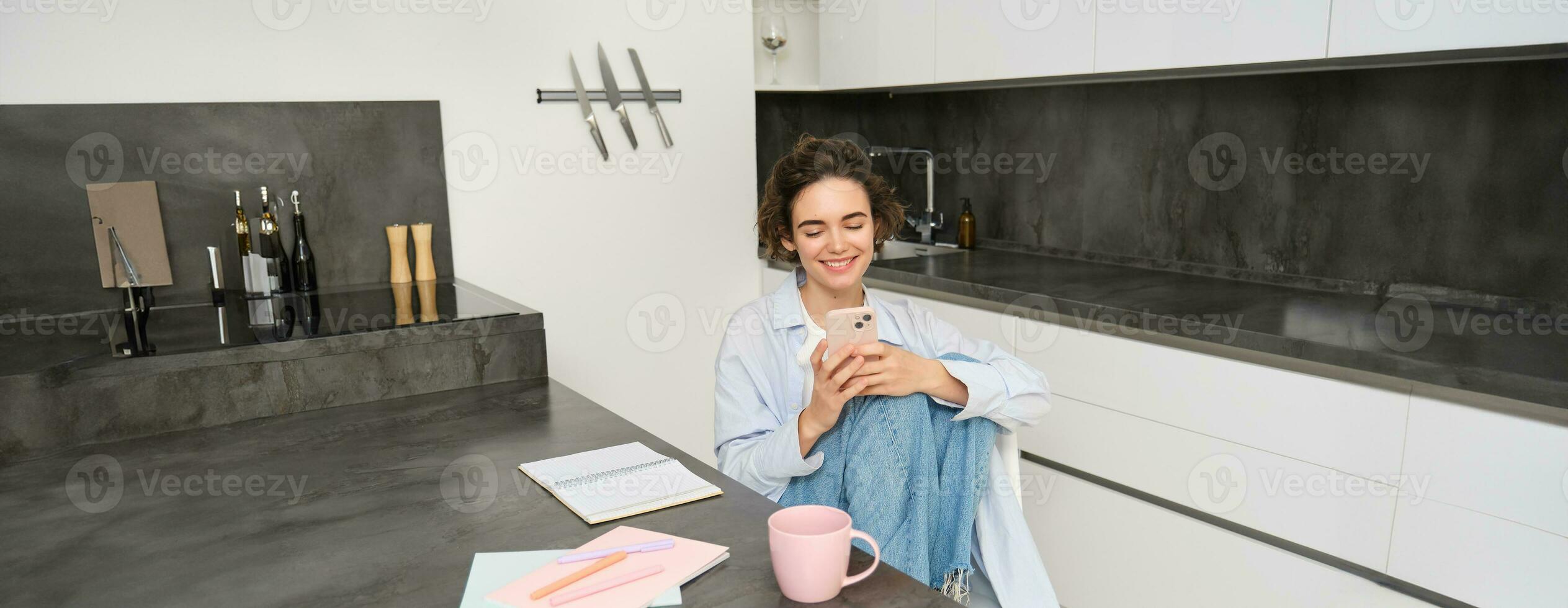 Young woman having break from work, sitting in kitchen and using smartphone, girl studies at home, sends a message on mobile phone photo